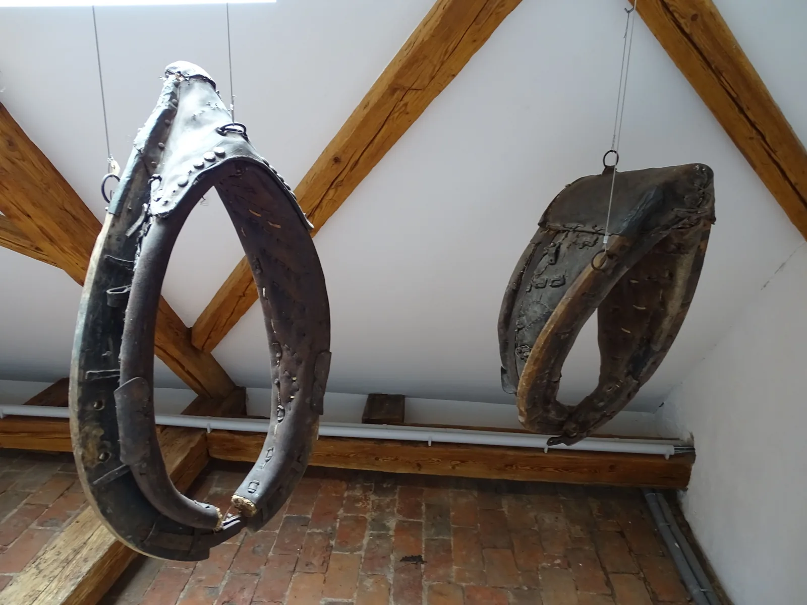 Zwei alte Kummete aus Holz und gepolstertem Leder hängen an Drähten unter einem Dachstuhl mit Holzbalken, das vordere zeigt die ovale Öffnung für den Hals.